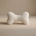 A cream bone-shaped pet toy with a small Woolmark black tag, resting on a neutral beige surface against a matching beige background.
