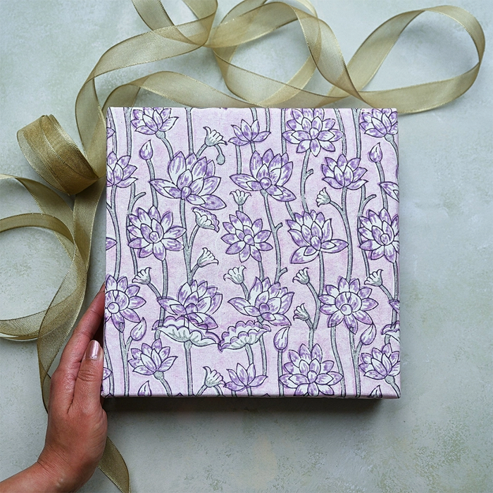 Handmade Indian Cotton Paper- Block Printed lotus floral pattern with lavender flowers and green stems used as gift wrap. Shown with a hand and green ribbon.