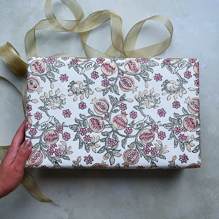 Block Printed pink and gold pomegranate pattern with  green leaves used to wrap a gift. Shown with a hand and gold ribbon.