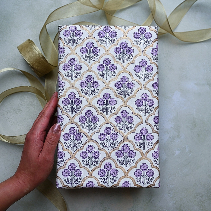 Block Printed lavender and green floral pattern with gold arches used to wrap a gift. Shown with a hand and gold ribbon.