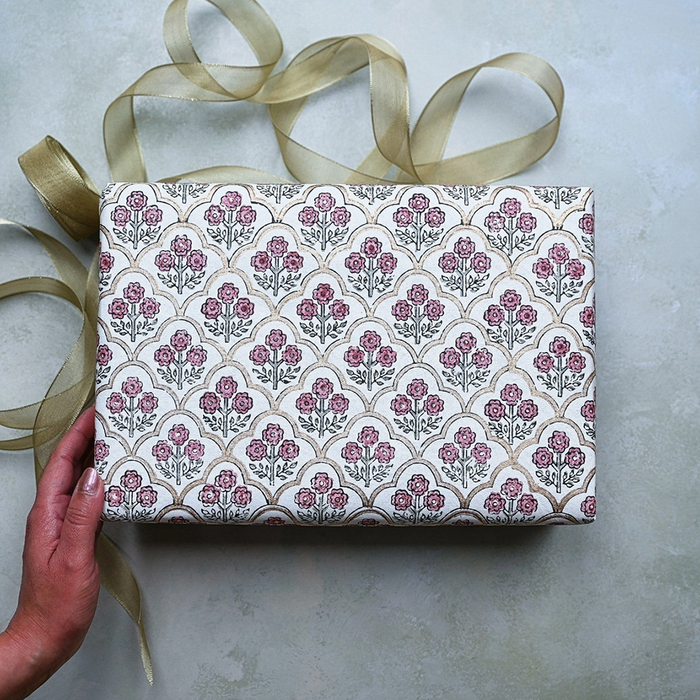 Block Printed pink and green floral pattern with gold arches used to wrap a gift. Shown with a hand and gold ribbon.