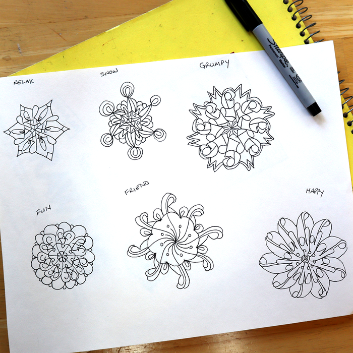 A sheet of white paper on a wooden table showing six black line-art mandala designs labeled Relax, Show, Grumpy, Fun, Friendly, and Happy. A yellow spiral notebook and a black Sharpie marker are partially visible around the paper.