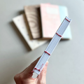 A hand holds a sewn text block of pages stitched with thin red thread, while three soft-focus notebook covers sit on a light surface in the background.