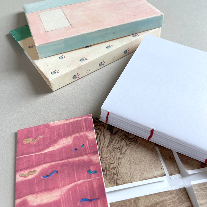 Handmade paper notebooks on a neutral beige surface. The stack shows three distinct covers: a top one with a textured pink and light blue design, a middle one with a cream base and small blue floral motifs, and a bottom one showing a pink textured cover with blue and gold abstract details with a white text block and red stitching