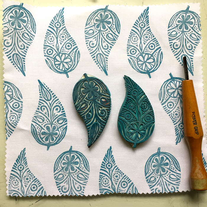 Two carved leaf-shaped carved blocks with teal printed patterns displayed on a white fabric with a wooden carving tool.