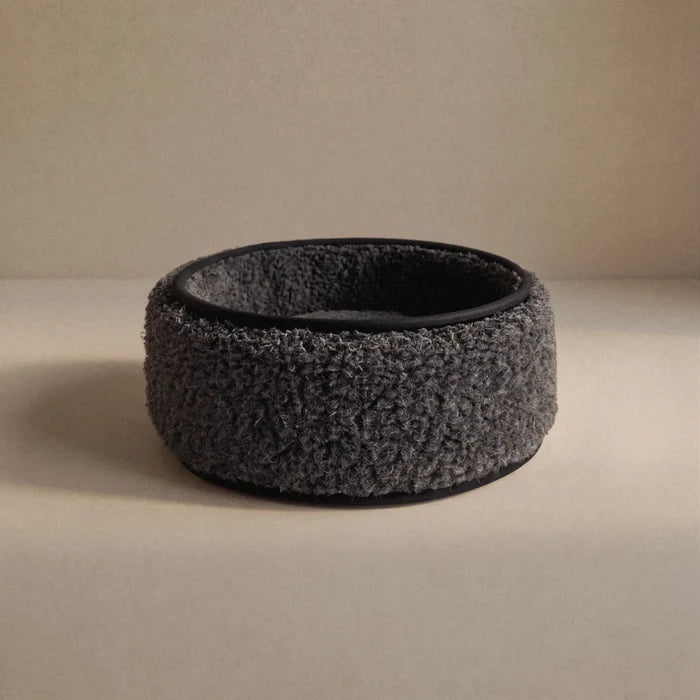 A dark wool sheep bouclé pet bed, with a black interior and base, photographed on a neutral beige surface and background.