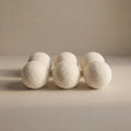 Six off-white wool balls arranged in two rows on a neutral beige surface against a matching beige background.
